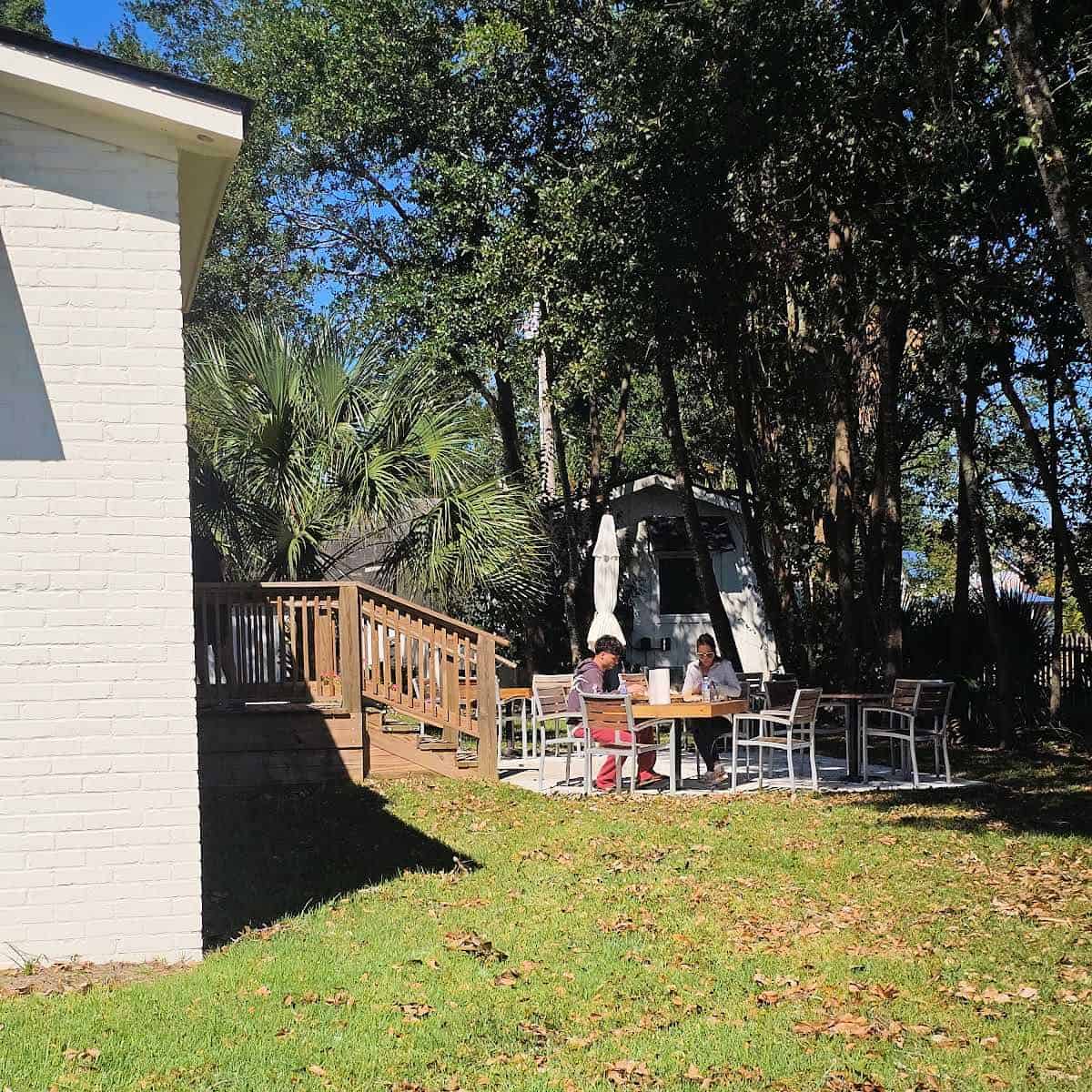 outdoor seating with tables and chairs next to a white brick building, trees along the side