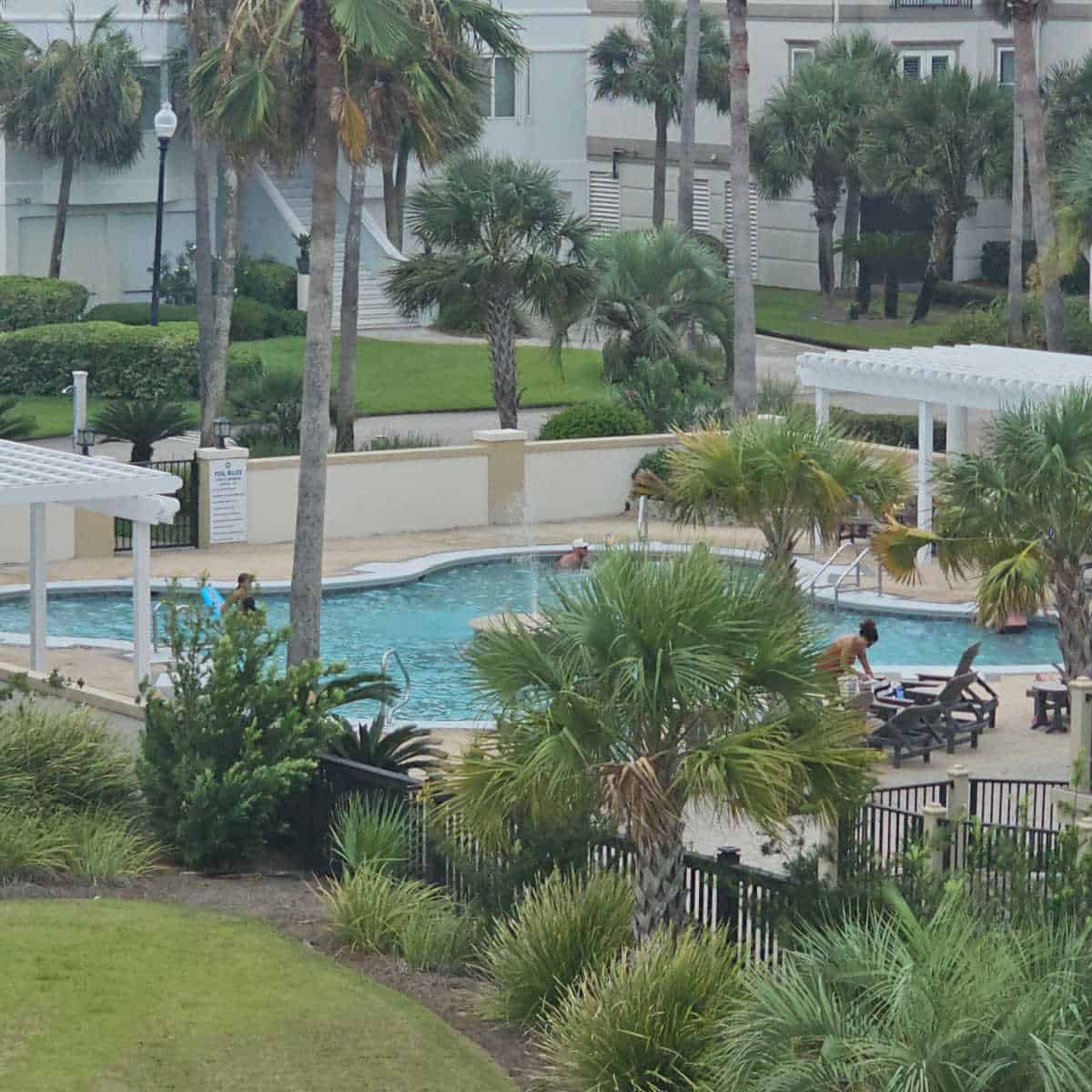 looking out towards the outdoor pool with people in the water, chairs on the pool deck and a dark fence around the pool area, palm trees outside the fence