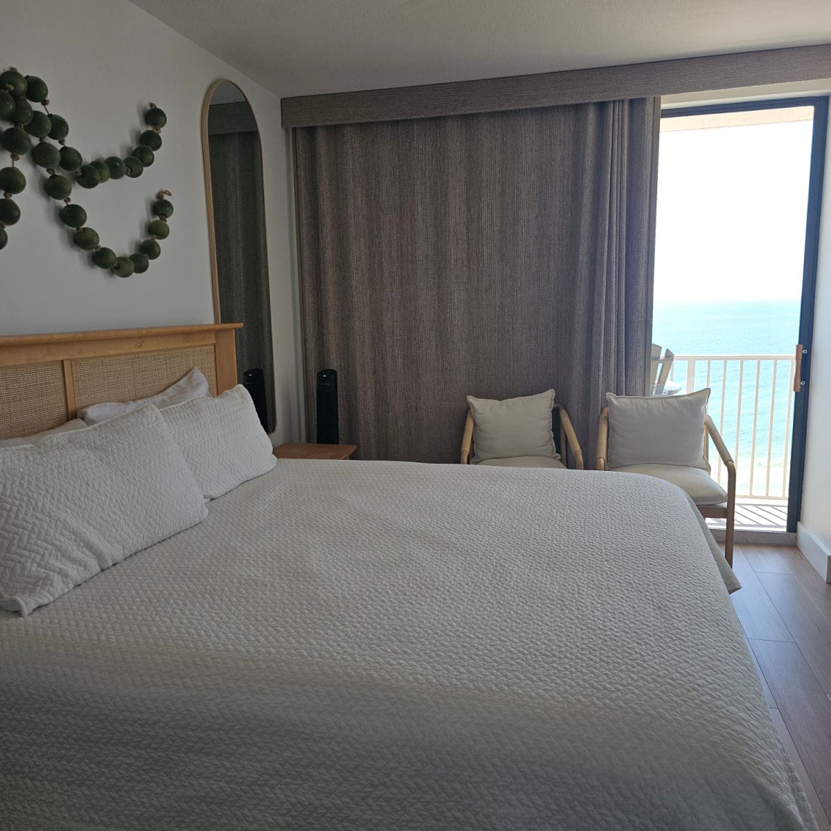 Bedroom with white linens, wooden headboard, green garland of balls on the wall, two chairs near the balcony access with the Gulf in the background