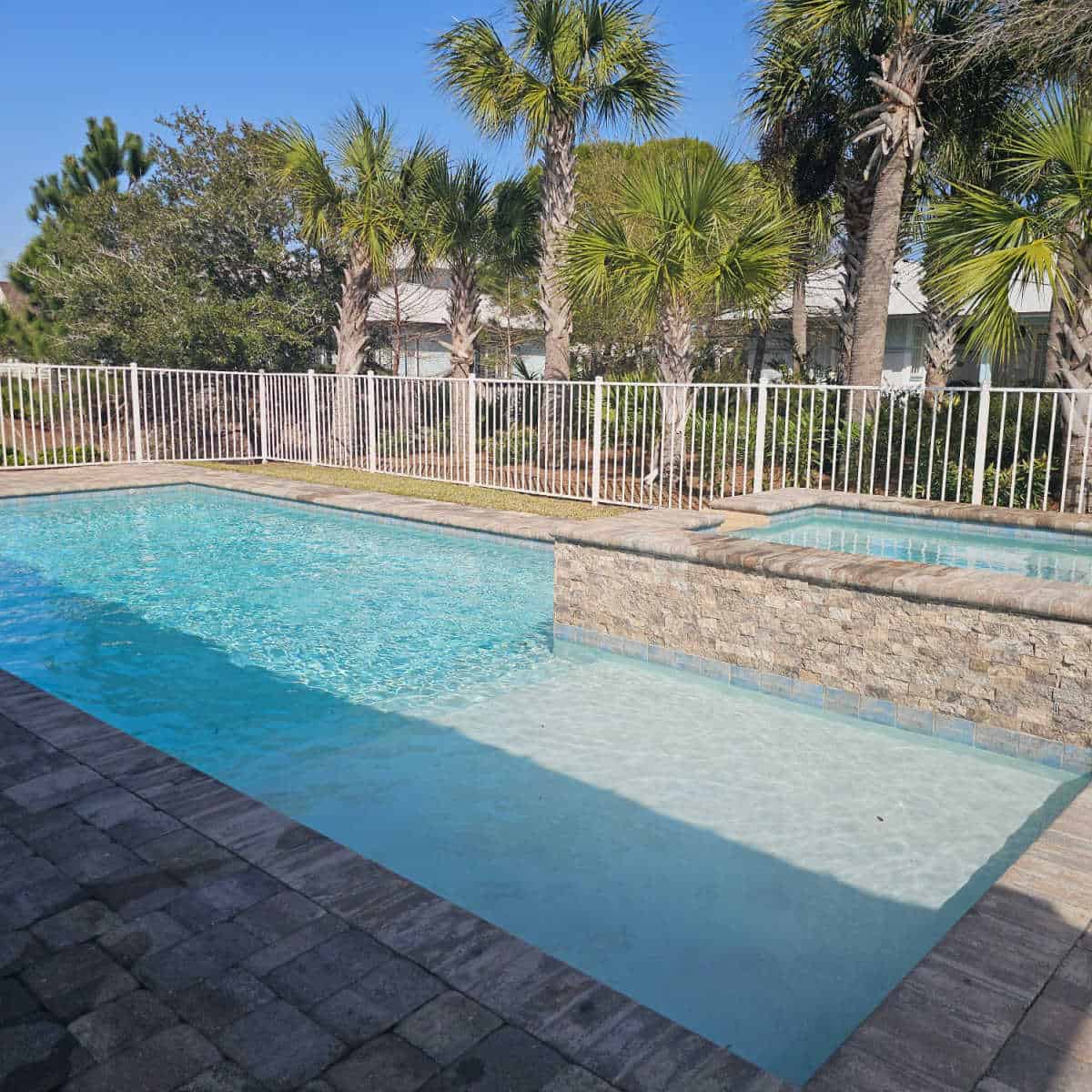 pool and hot tub behind a white fence with palm trees behind it
