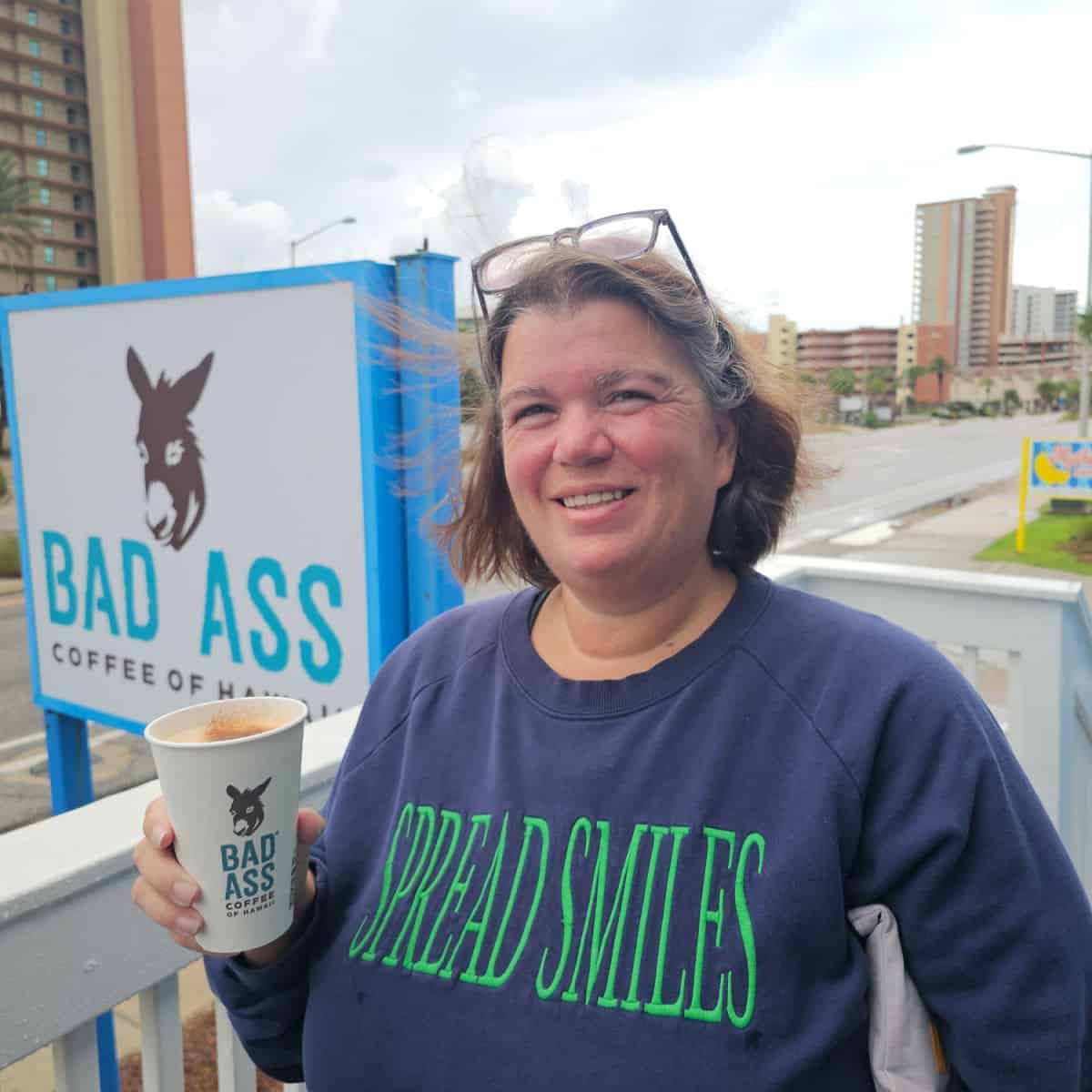 Tammilee holding a Bad Ass Coffee of Hawaii cup next ot the bad ass coffee sign with condo buildings in the background