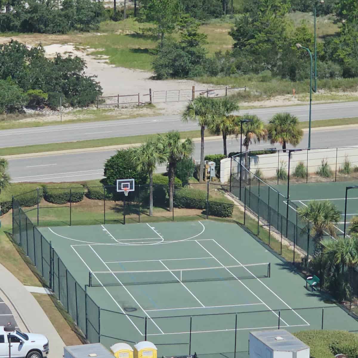 looking down to a tennis court with a basketball hoop at one end, surrounded by a tall fence, Perdido Beach Blvd behind the tennis court