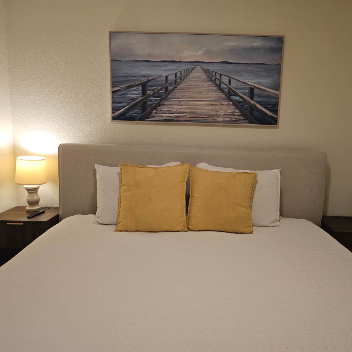 Bedroom with white linens and two yellow pillows, nightstand with a lamp on it, dock painting over the bed