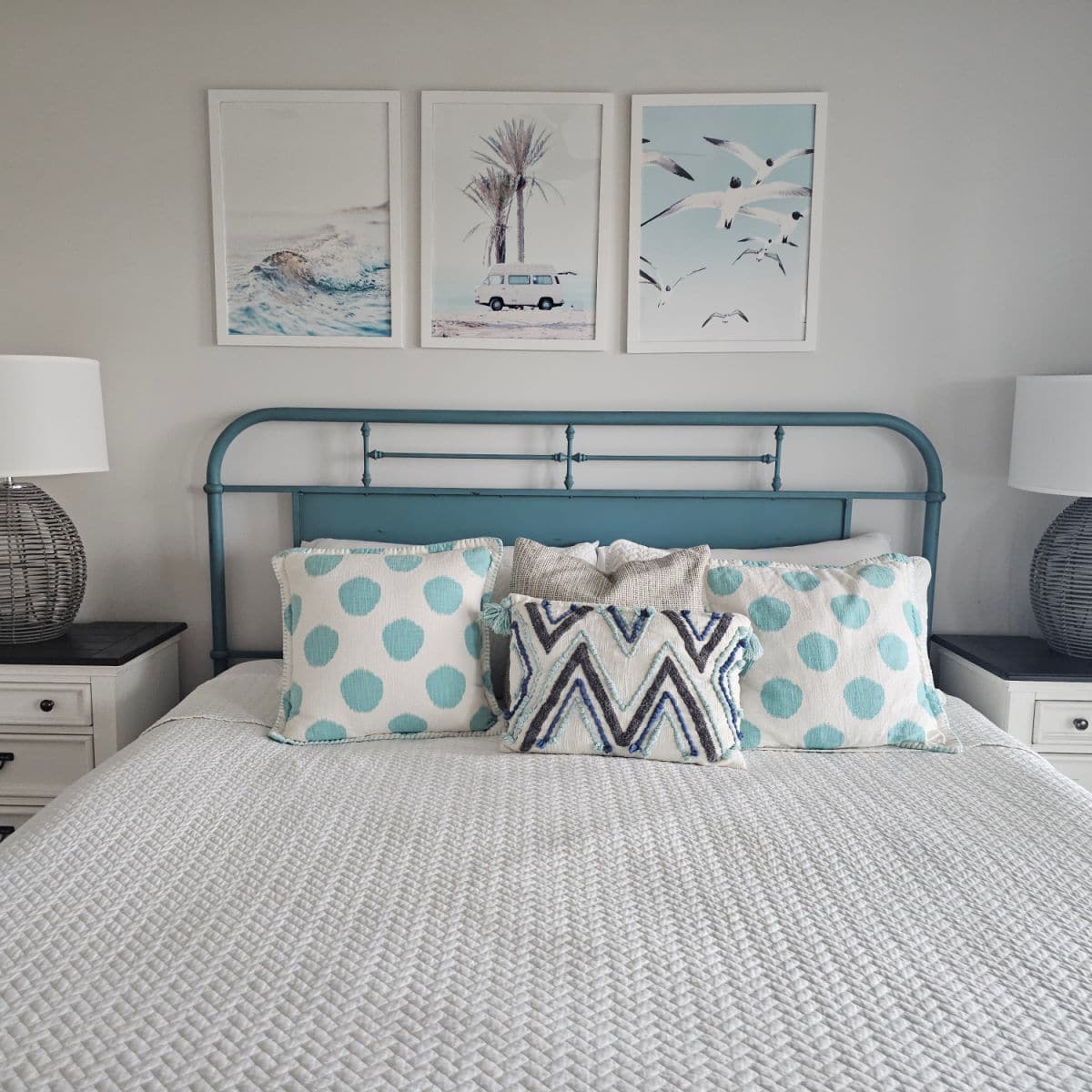 Bedroom with coastal art on the wall, teal bedframe with white linens and colorful pillows