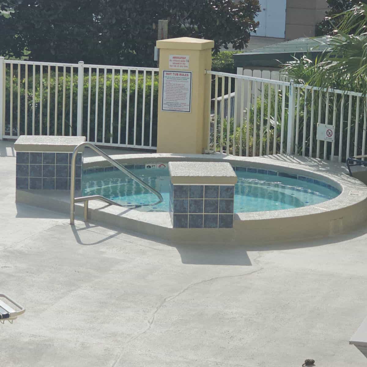 hot tub with a handrail going into the hot tub, pool rules on a column next to the white fence