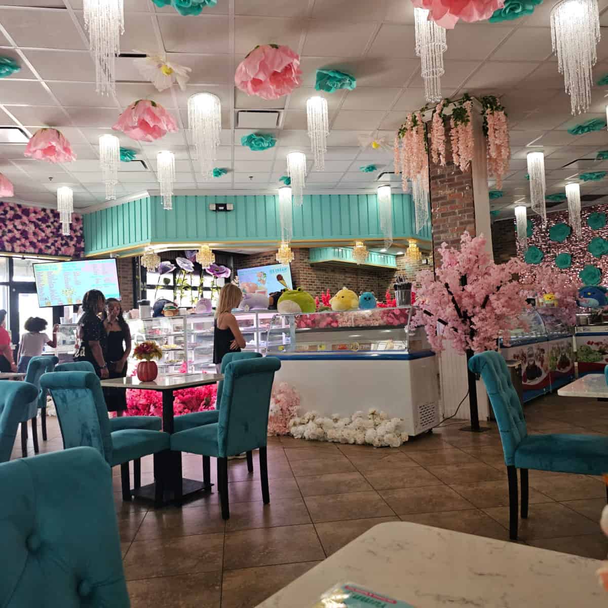 looking towards the order counter with flowers and lights hanging from the ceiling, blue chairs at tables, people standing next to a bakery case