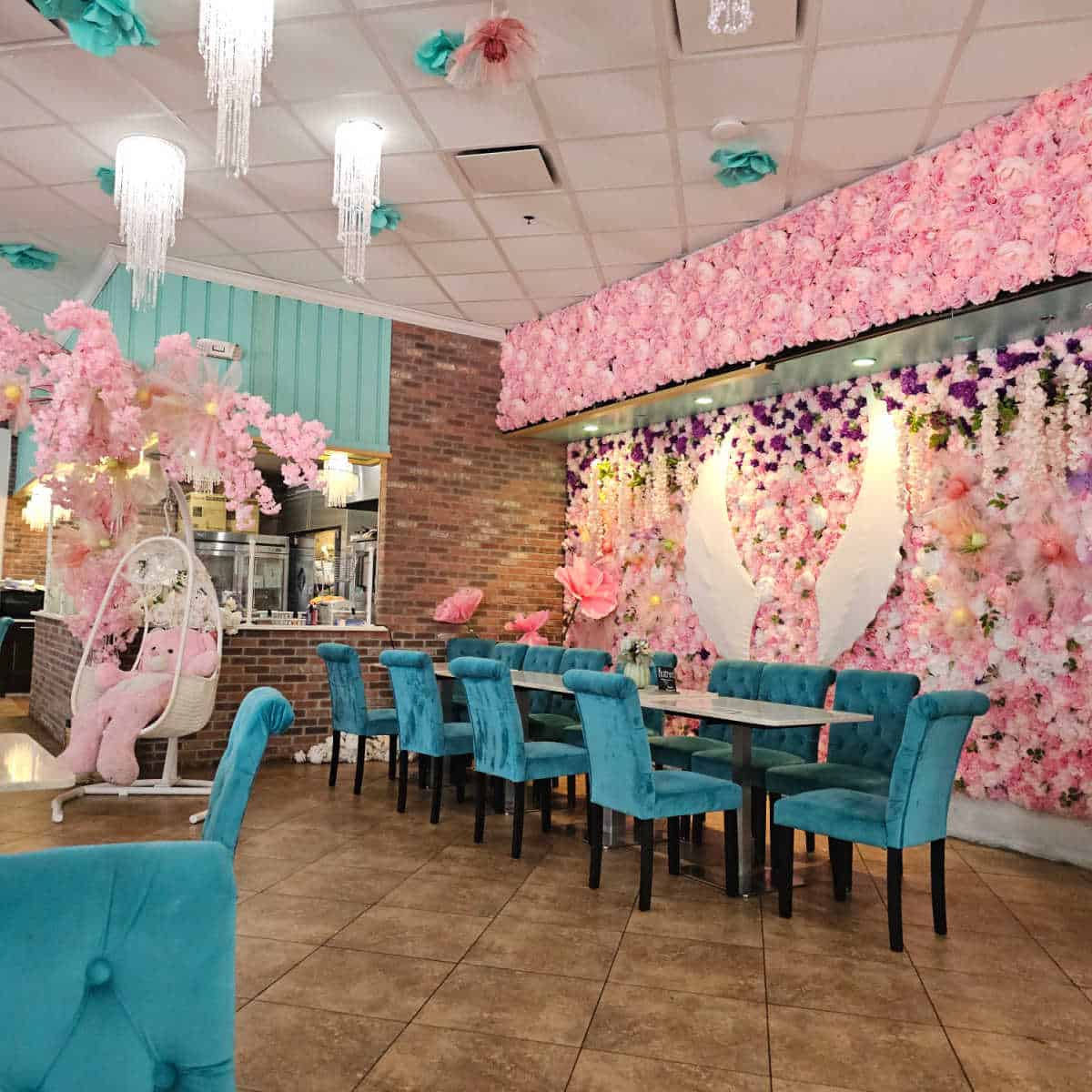 wall covered in flowers and a pair of angel wings, long table with blue chairs, teddy bear in a swing chair, flowers and lights hanging from the ceiling