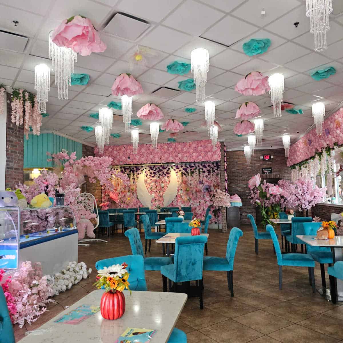 super colorful restaurant interior with flowers and lights hanging from the ceiling, blue chairs and white tables, large angel wings on the back wall with flowers