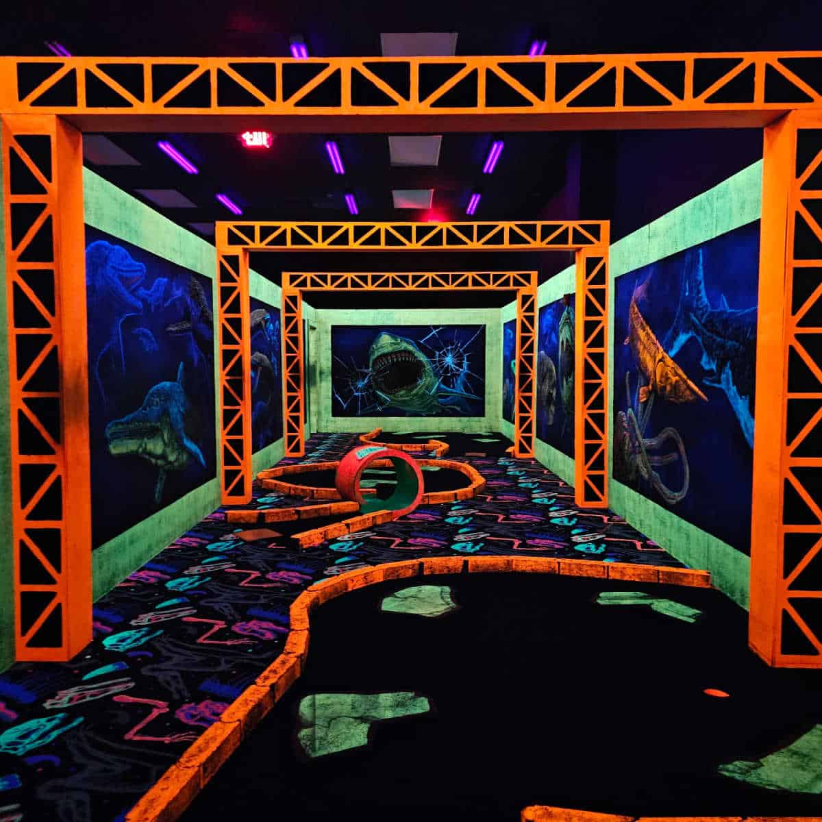 looking down the indoor mini golf course with orange walk throughs glowing and a shark are the end of the room. mini golf courses in the middle, glowing with blacklight and neon