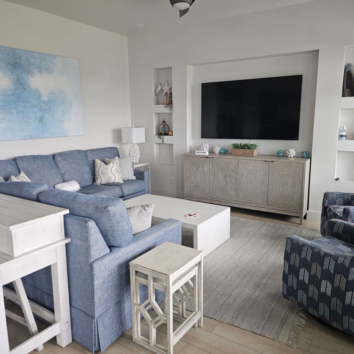 Living room area with blue couches, white tables, flat screen tv on the wall over a wooden consol, chairs on the side