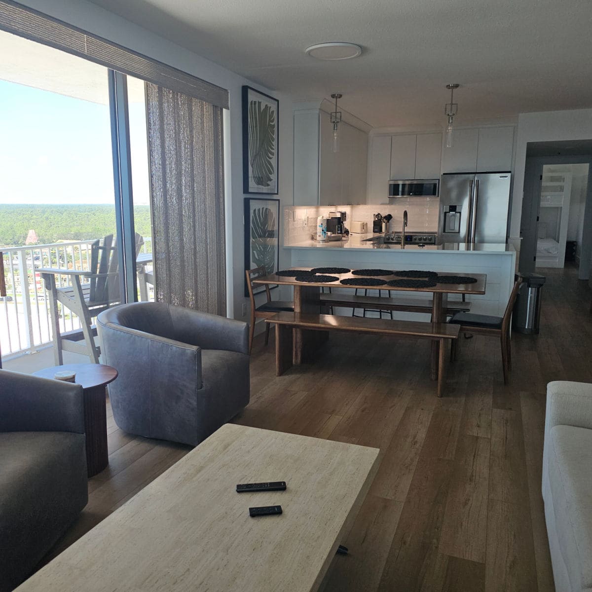looking across the living room the kitchen an a bedroom in the background. Two leather chairs with a table near them, dining room table with palm leaf art on the wall, kitchen with white cabinets and stainless steel appliance