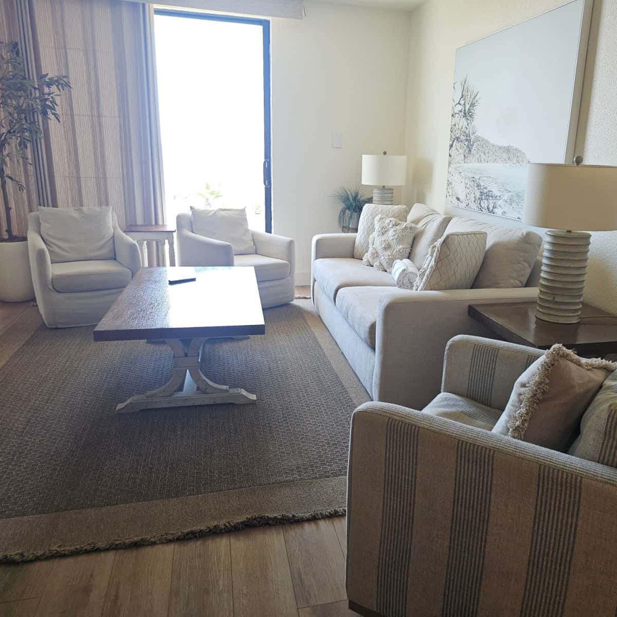 Living room with a beige couch and two chairs, a striped chair, table and lamps, with a fake olive tree in the corner