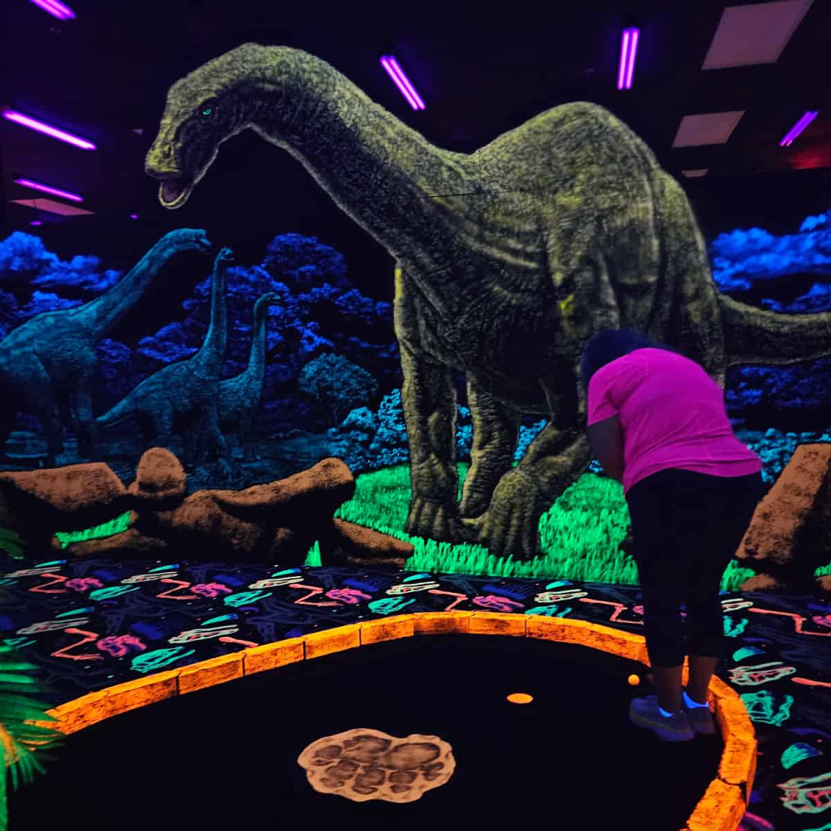 Large dinosaur painting glowing over a female playing mini golf in a neon course