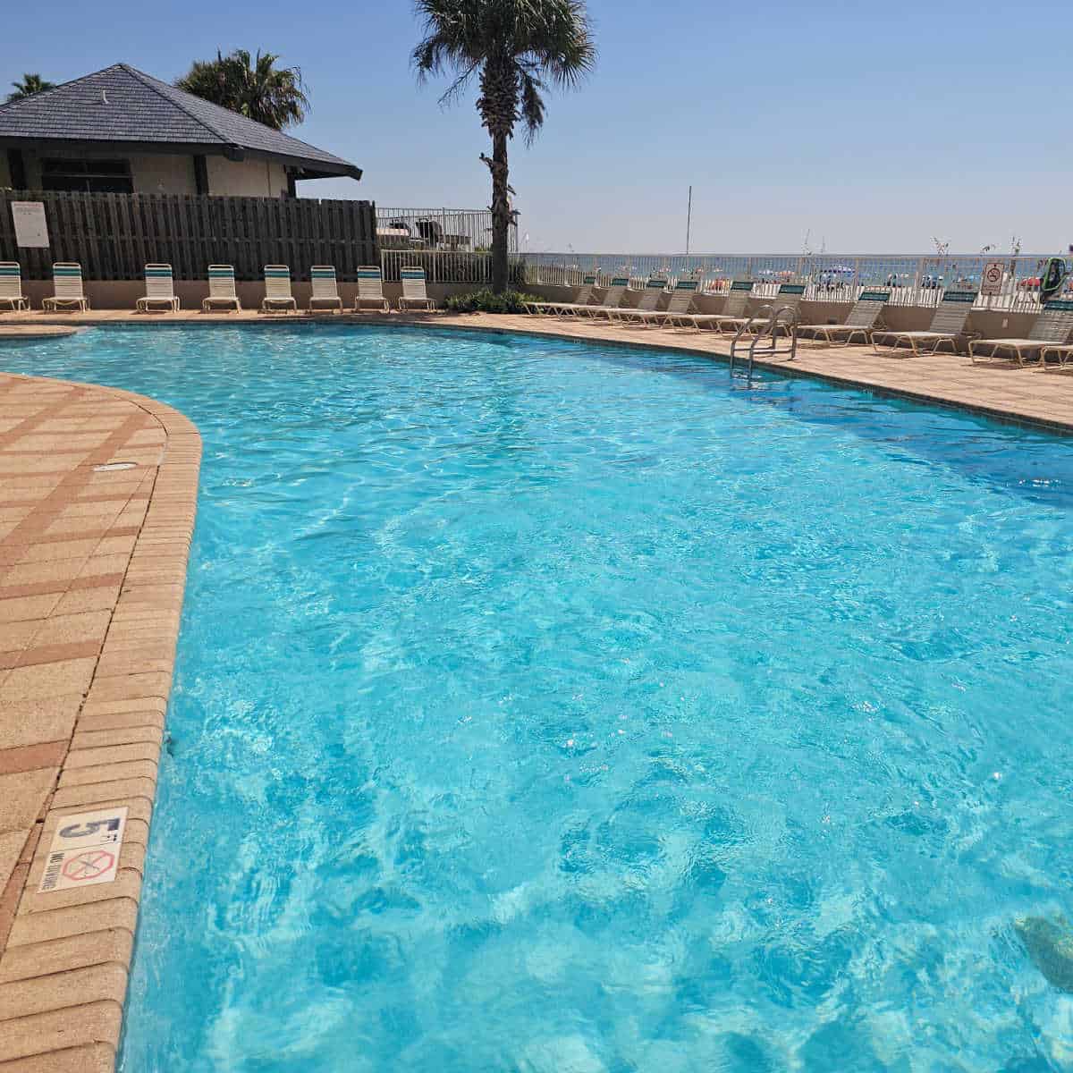 Outdoor pool sparkling in the sun with lounge chairs around the fenced in pool deck, beach views in the background