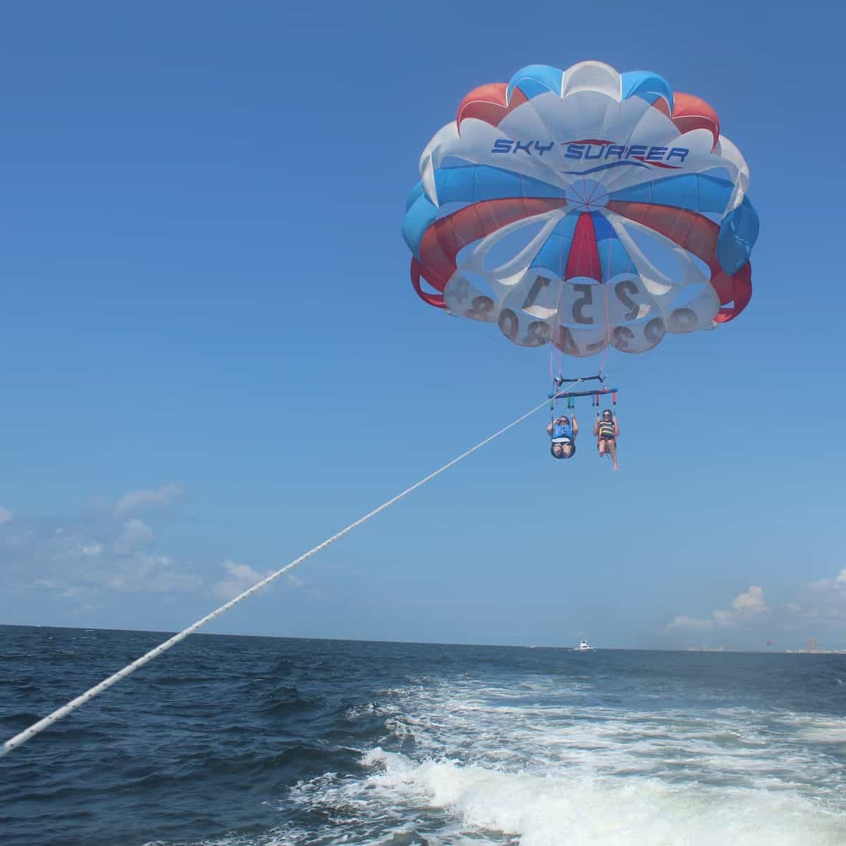 Red white and blue parachute with Sky Surfer printed on it, Tammilee and Friend parasailing with a rope to the boat