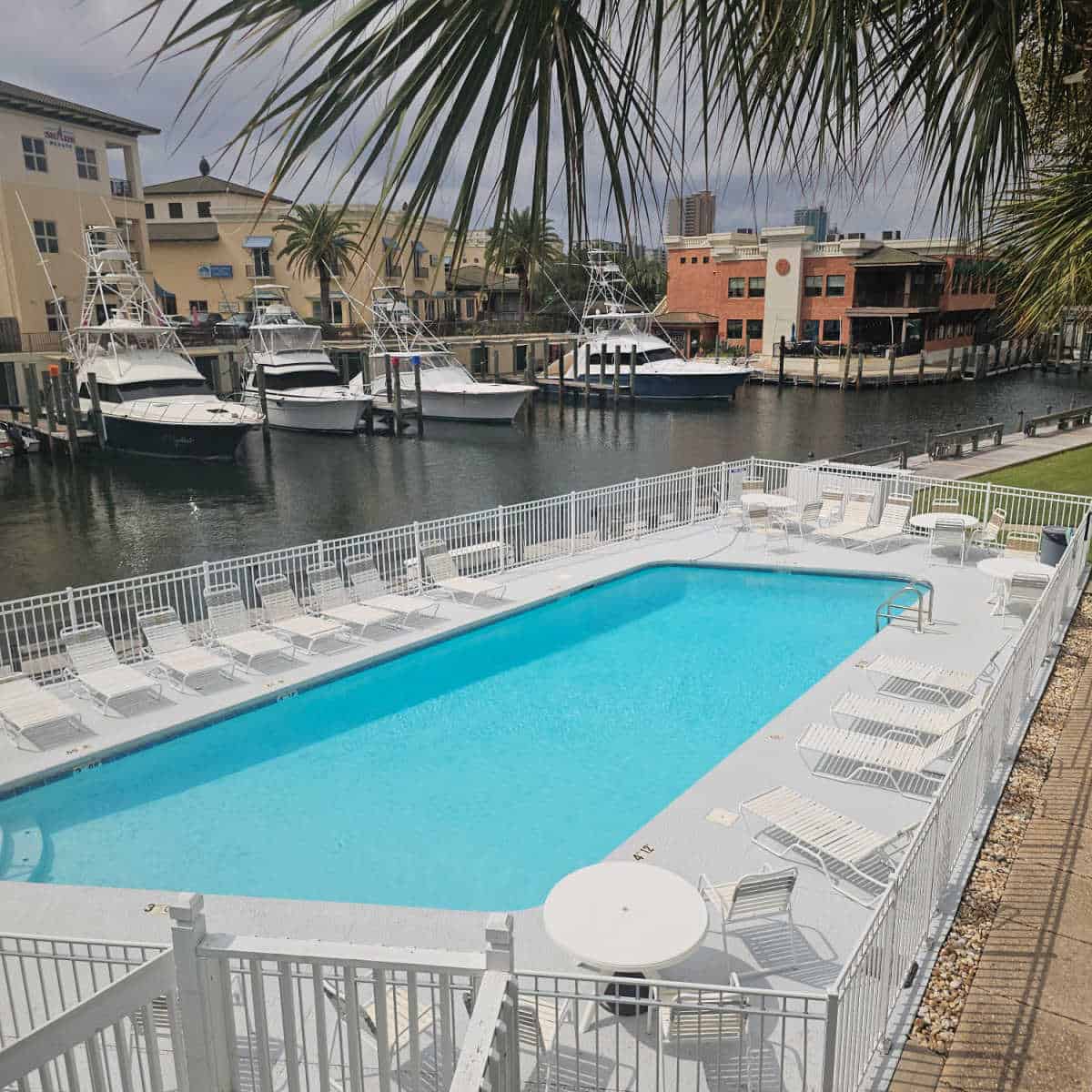 looking over an outdoor pool with white lounge chairs and a white fence around the pool area, water with boats docks and buildings behind them.