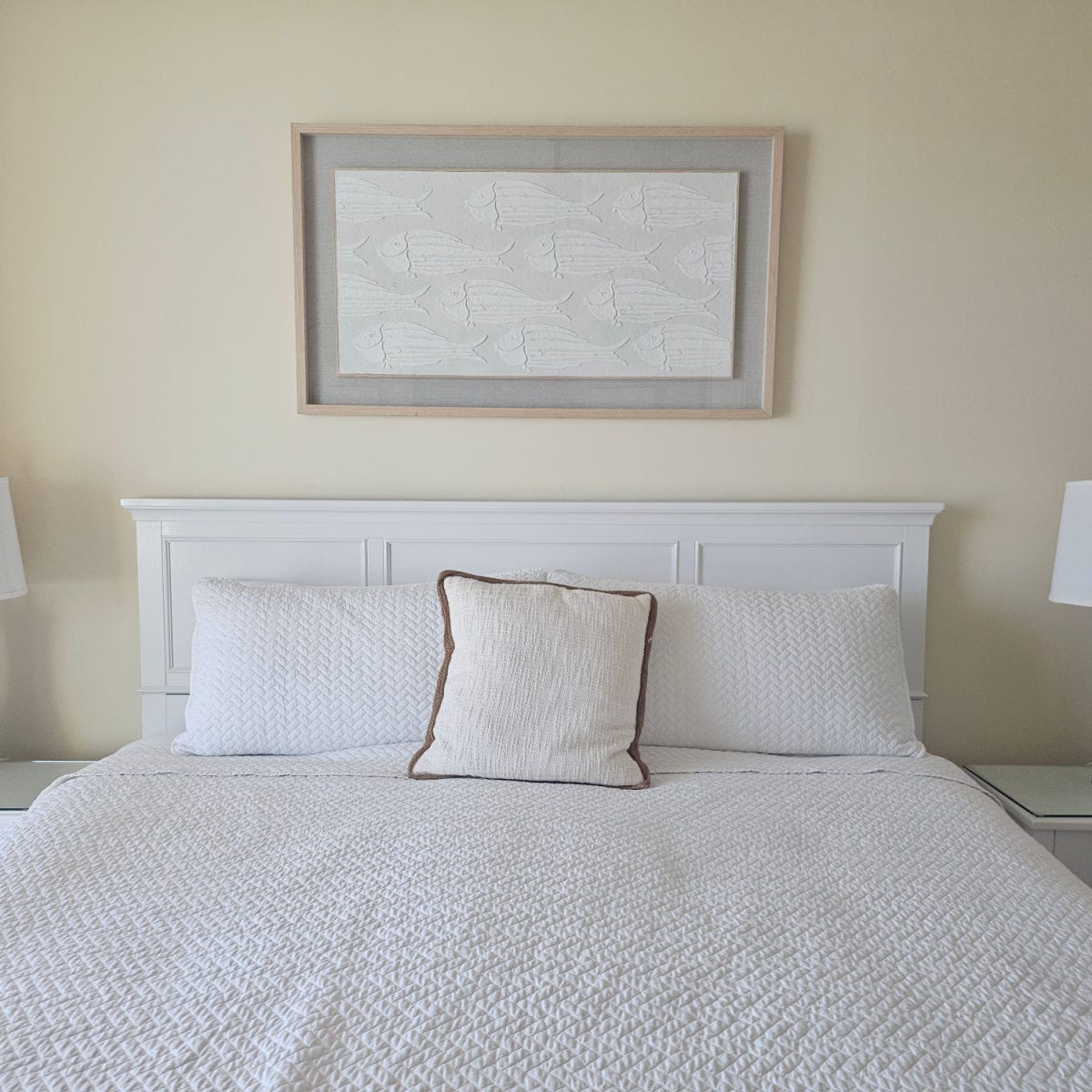 bedroom with light yellow walls, fish art on the wall, bed with white headboard and white linens, portion of white lamps on the sides of the bed