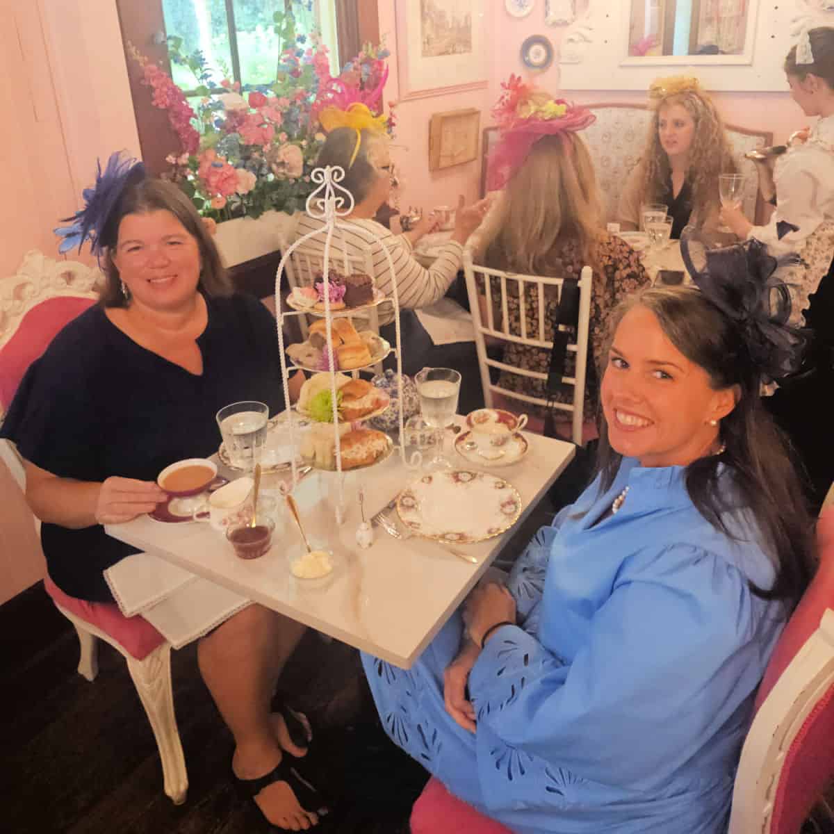 Tammilee and eva sitting at a table with a tiered serving tray, cups of tea, and a group of women sitting at a table behind them