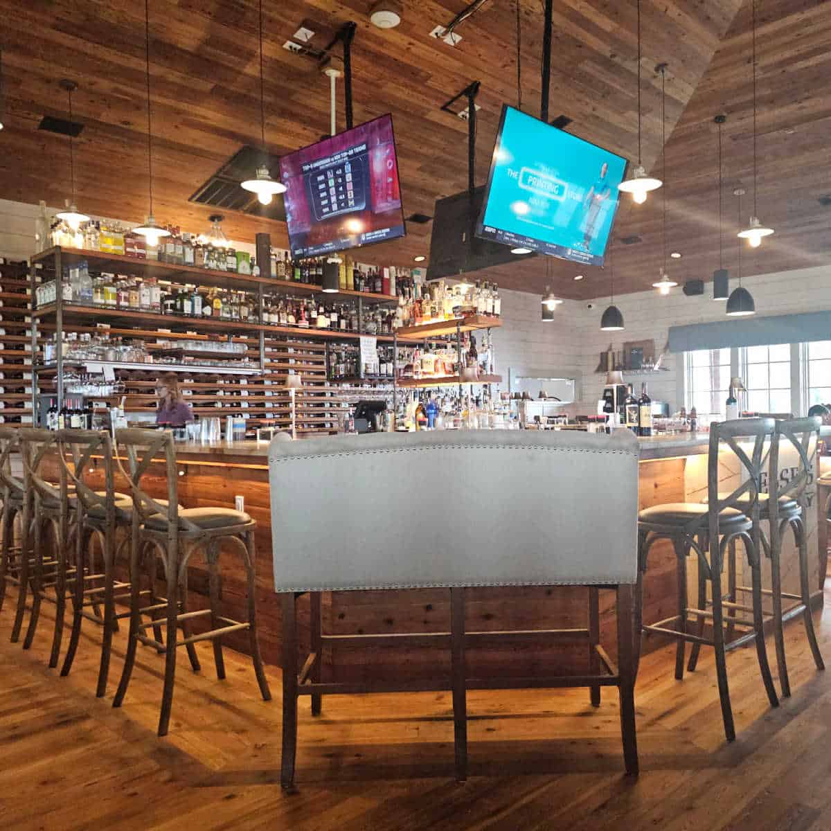 Bar area with a large raised bench and bar stools wrapped around the bar, Shelfs of bottles and glasses behind the bar, tvs hanging from the ceiling