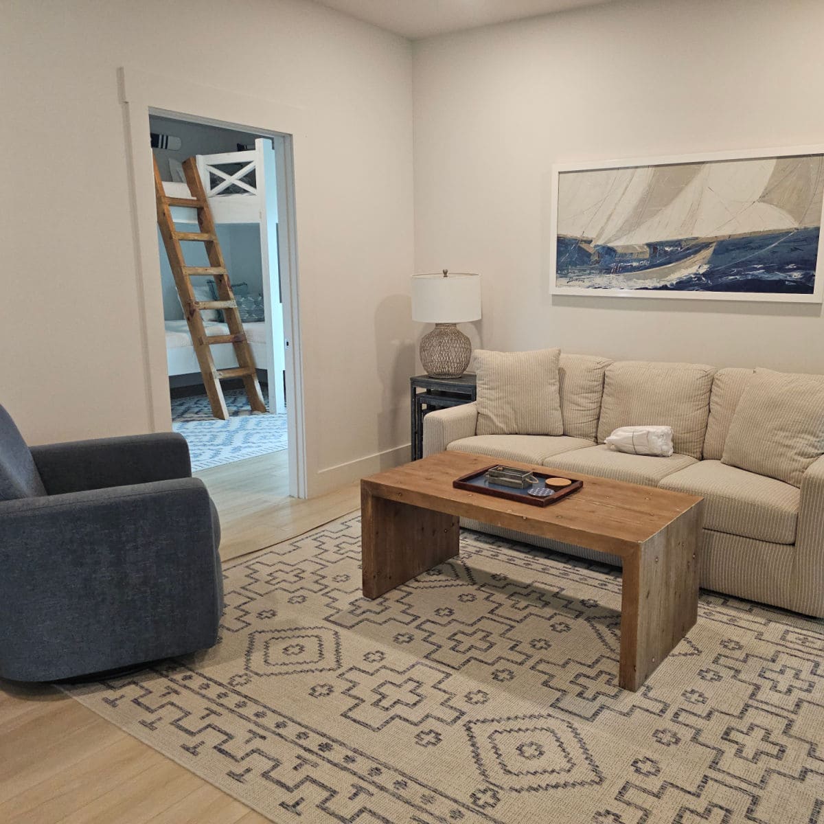 Sitting area with a cream colored couch, wooden table, sailboat art above the couch, and blue chair, looking through a doorway to a set of bunkbeds